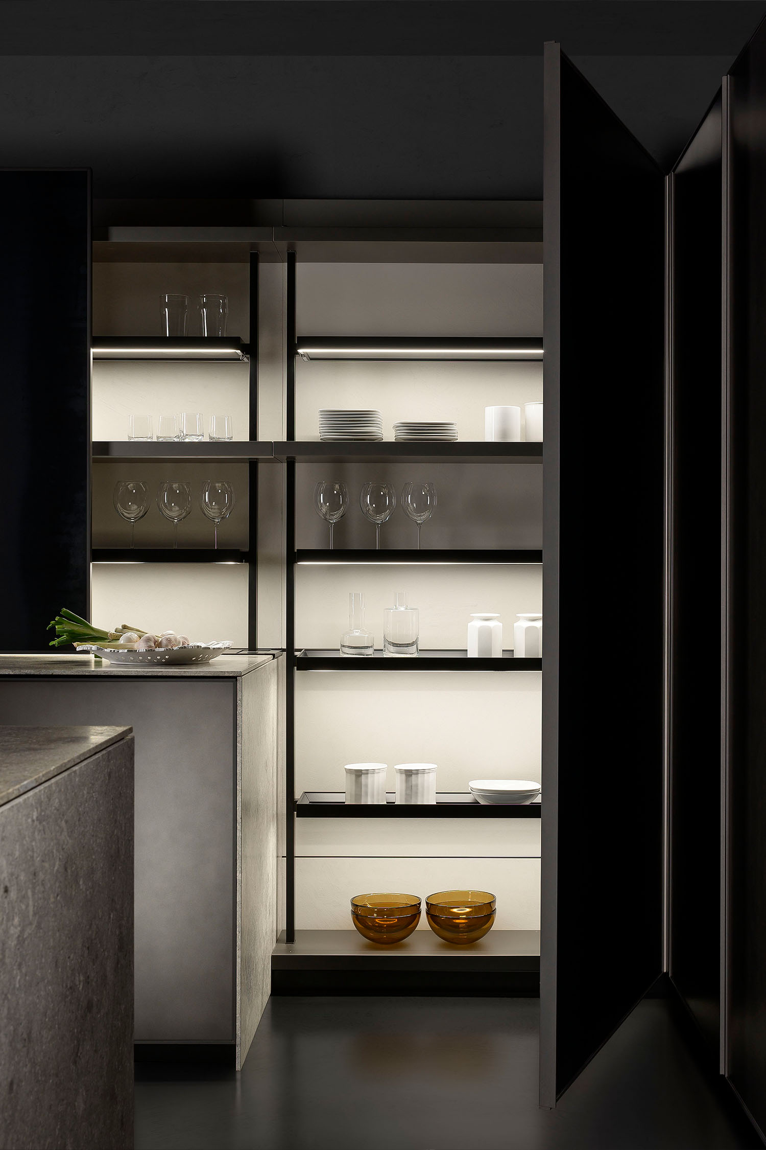 A detail shot of a modern kitchen countertop with a wooden, round bowl and a small plant. The countertop is a light, stone-like material, and the background is a smooth, grey wall.