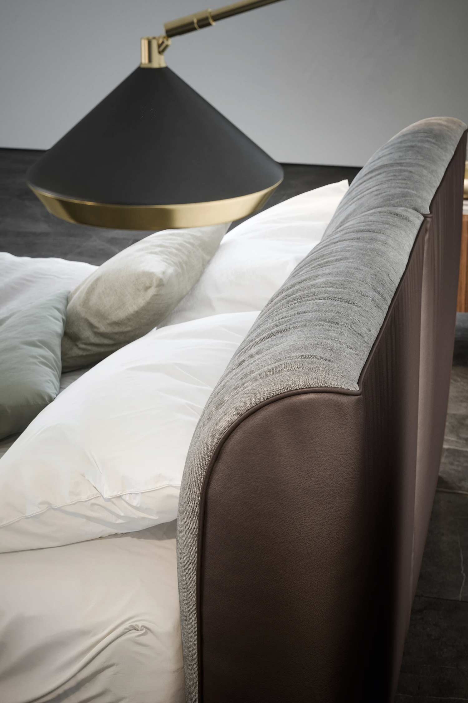 A close-up detail shot of a two-toned upholstered headboard. The inner, slightly ribbed section is grey fabric, and the outer, curved border is smooth brown leather or faux leather. White pillows rest against the headboard, and a large, black and brass cone-shaped lamp is visible in the background.
