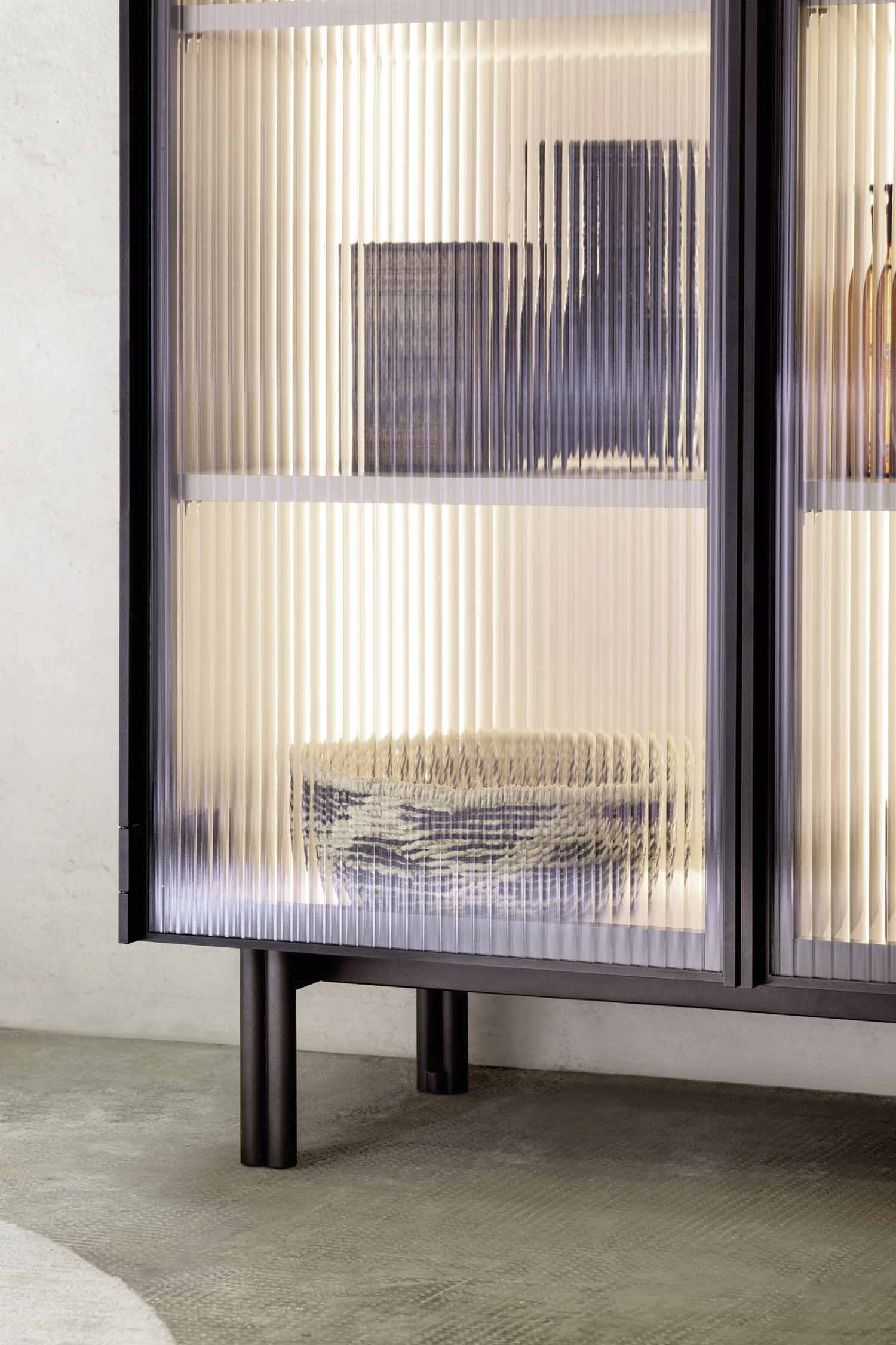 A close-up shot of the lower section of a modern display cabinet. The image focuses on the reeded glass doors and the light glowing from inside. A dark-colored woven basket and other items are visible on the shelves. The cabinet is supported by simple, dark metal legs.