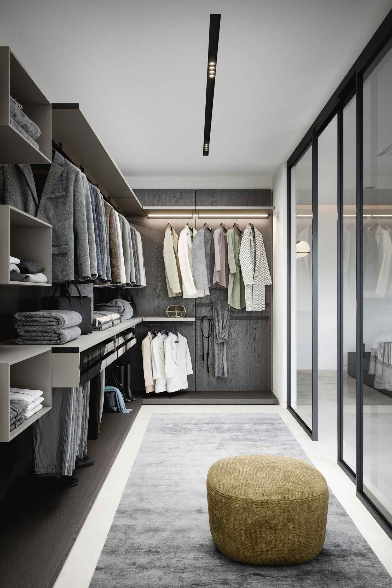 An interior view of a contemporary walk-in closet. The space features dark wood grain back panels and light gray shelves. A variety of clothes are hanging on racks and folded on shelves. A gray textured rug is on the floor, and a mustard-yellow ottoman sits in the foreground. Glass sliding doors are visible on the right, leading to another room.