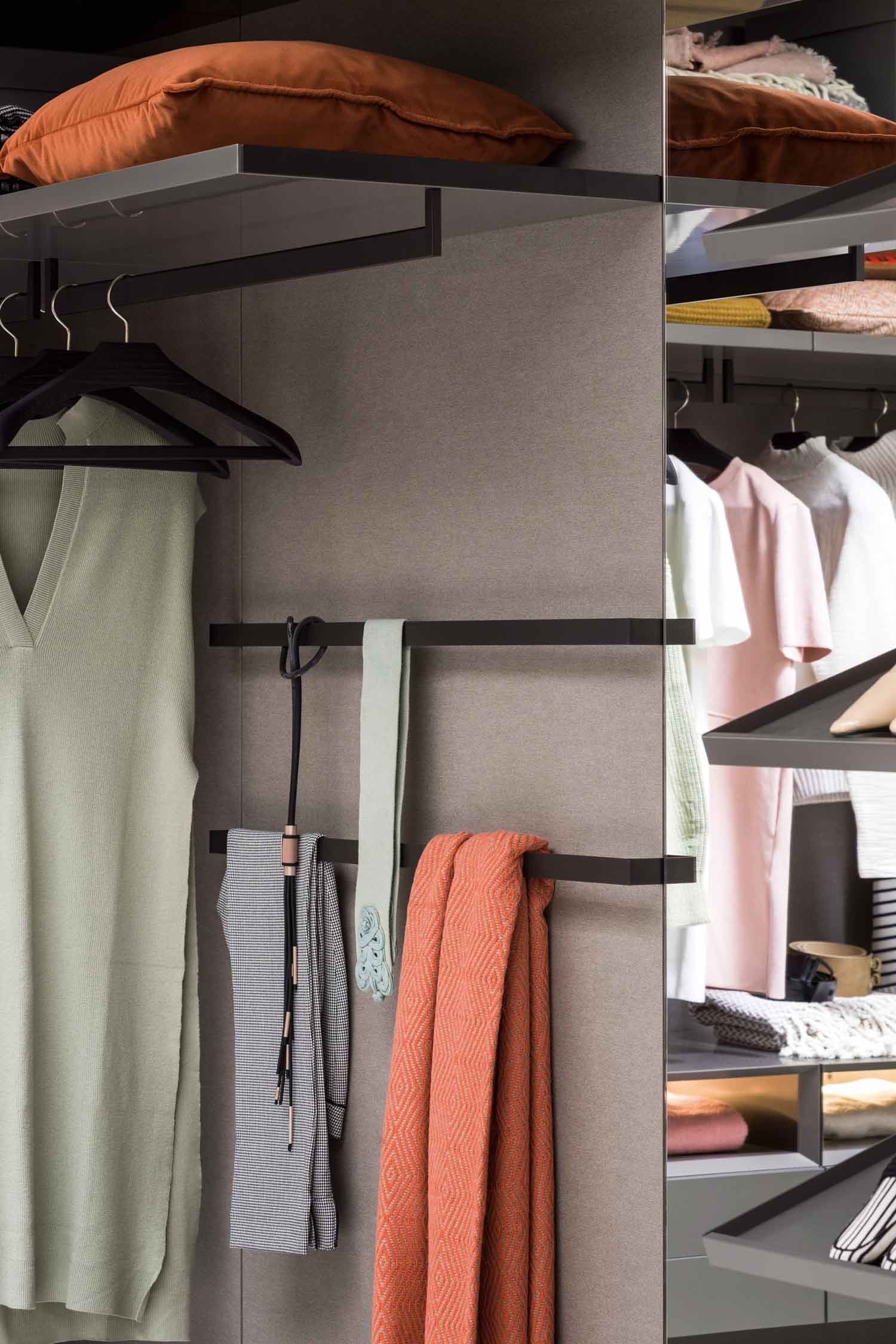 A close-up shot of a wardrobe's interior, focusing on clothing and accessories. A light-colored top hangs on a black hanger, and below it, a pair of patterned pants, a light-green belt or tie, and a folded orange scarf are hanging. The background is a textured gray material, and a reflection in a mirrored surface shows a shelf with more clothing.
