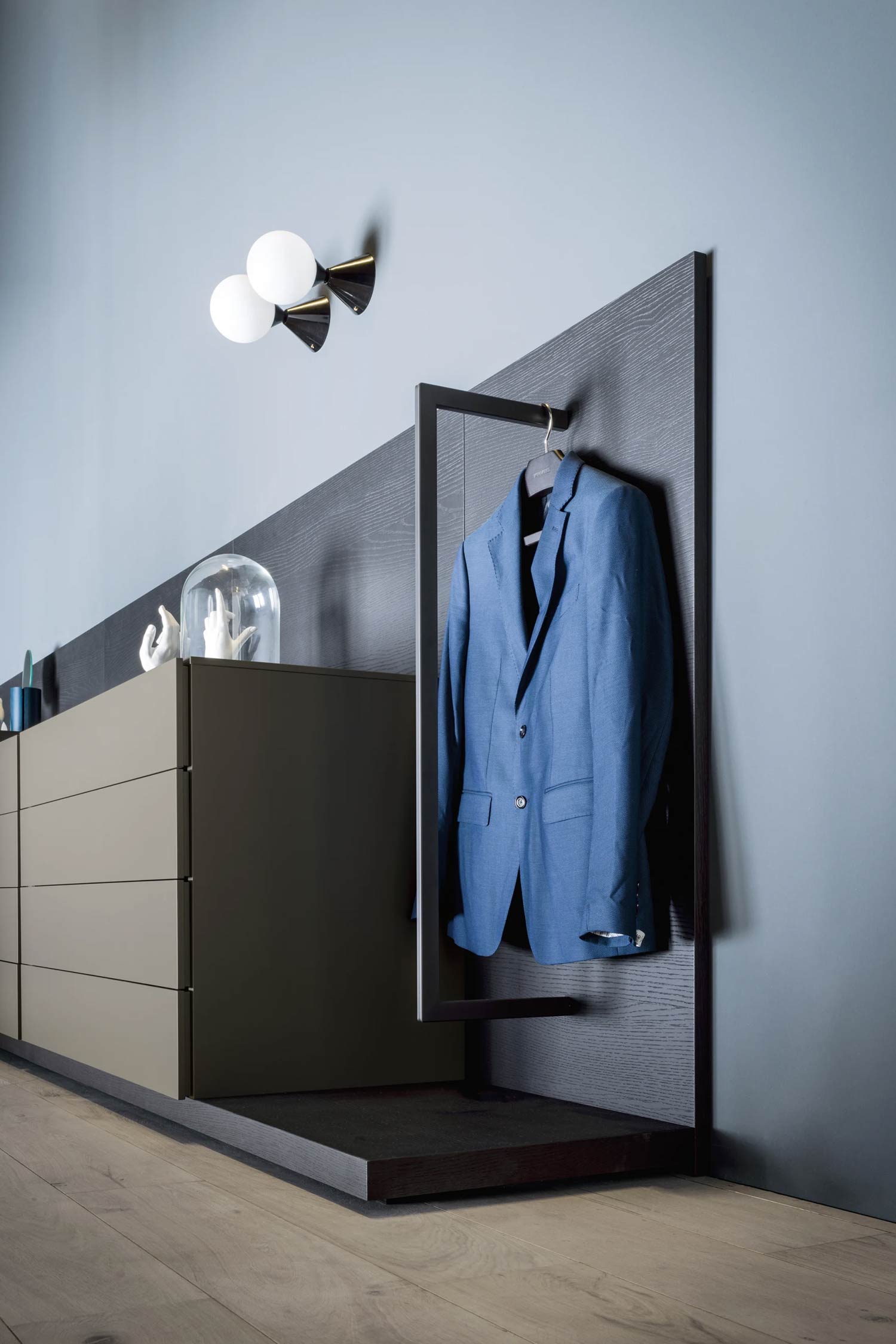 A modern closet or dressing area detail featuring a low, long drawer unit in a muted taupe color next to a vertical dark wood display panel with an integrated black metal clothes rack holding a bright blue suit jacket. White and black wall sconces are mounted above on a blue wall.