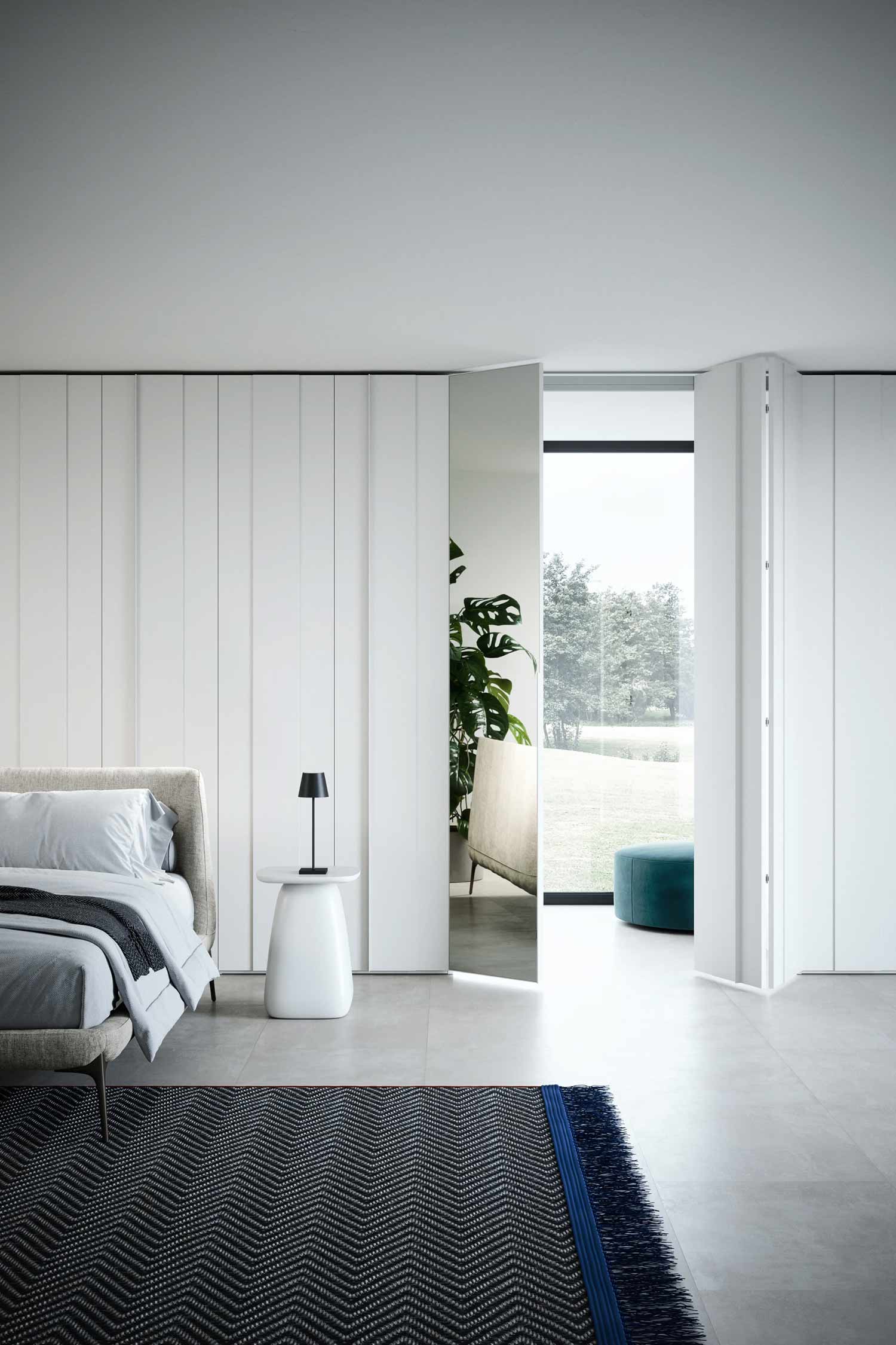 A bright, minimalist bedroom. A beige upholstered bed with gray bedding is on the left. The far wall is lined with a seamless, floor-to-ceiling white hinged wardrobe with a pleated design. One section of the wardrobe is open, revealing a room with a large window that looks out onto a grassy landscape. A lounge chair and a small teal ottoman are visible in the room. A white rug with a blue and black pattern is on the floor in front of the bed.