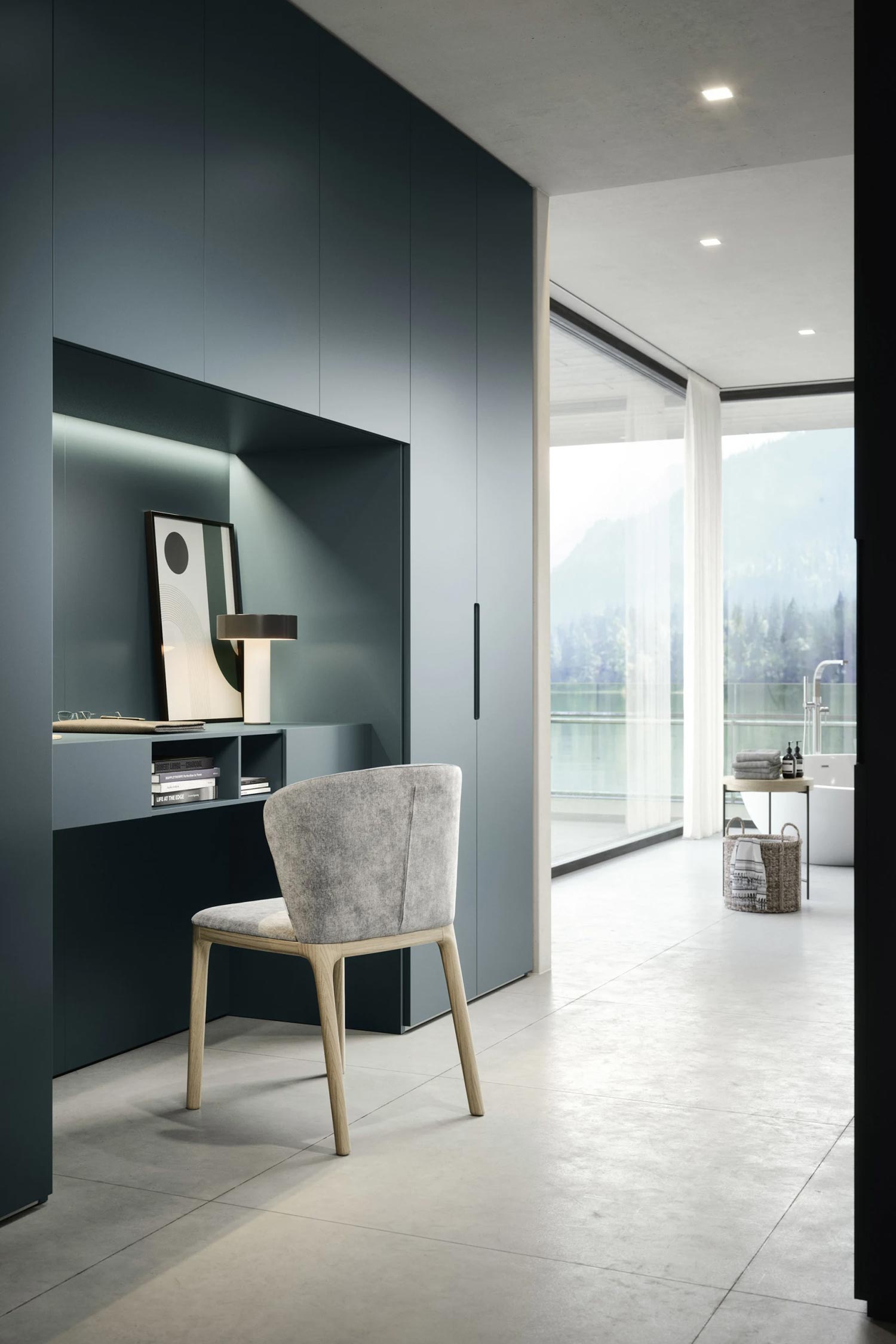 This image shows an integrated desk and workspace built into the side of a dark, matte Vico wardrobe. The desk features a lamp, a framed picture, and a chair. In the background, a bathroom with a free-standing tub is visible through an open doorway, with a large window overlooking a forest and lake.